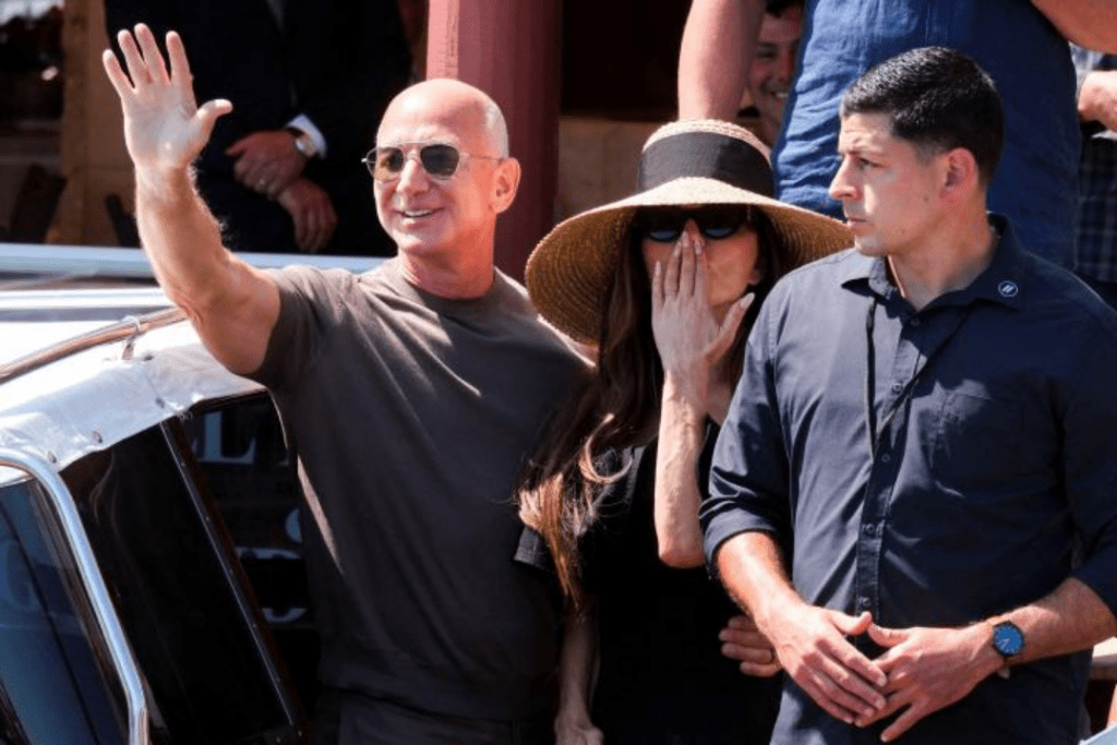 Jeff Bezos waves cheerfully beside Lauren Sanchez, who wears a wide straw hat, as they stand outdoors with another guest at what appears to be a social gathering.