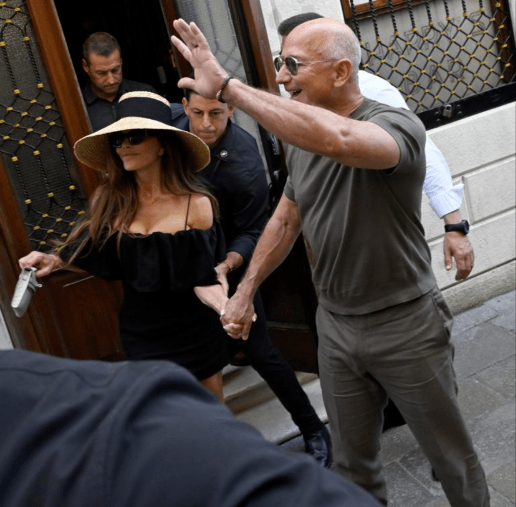 Jeff Bezos and Lauren Sanchez exit a building hand in hand, surrounded by security. Bezos wears a grey outfit and raises one hand in acknowledgment, while Sanchez stuns in a black off-shoulder dress and wide-brimmed hat.