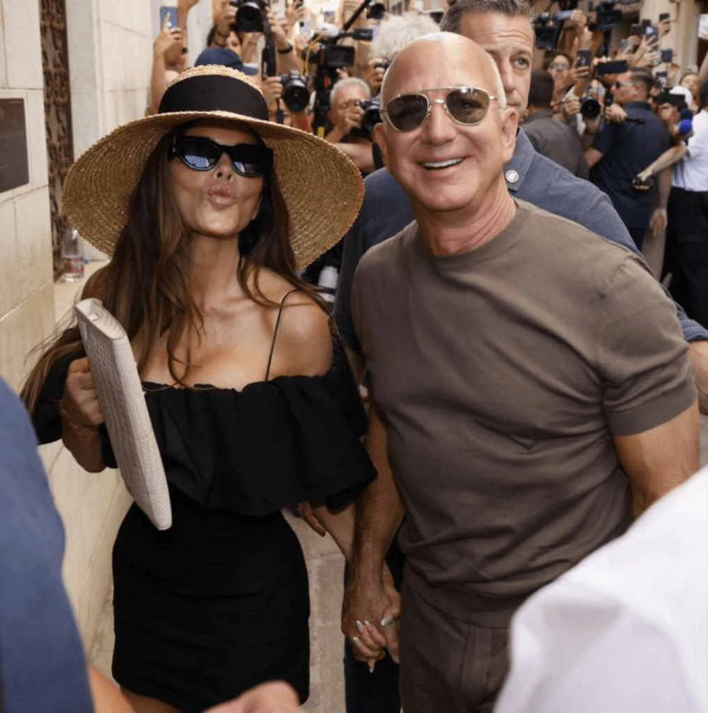 Jeff Bezos and Lauren Sanchez walk hand in hand through a crowd of photographers and onlookers. Sanchez wears a black off-the-shoulder dress with a wide-brimmed straw hat and carries a beige clutch, while Bezos is dressed casually in a brown t-shirt and pants.