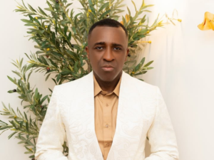 Frank Edoho stands confidently in a white patterned blazer over a tan shirt, framed by a backdrop of lush green foliage that enhances his polished and stylish presence.