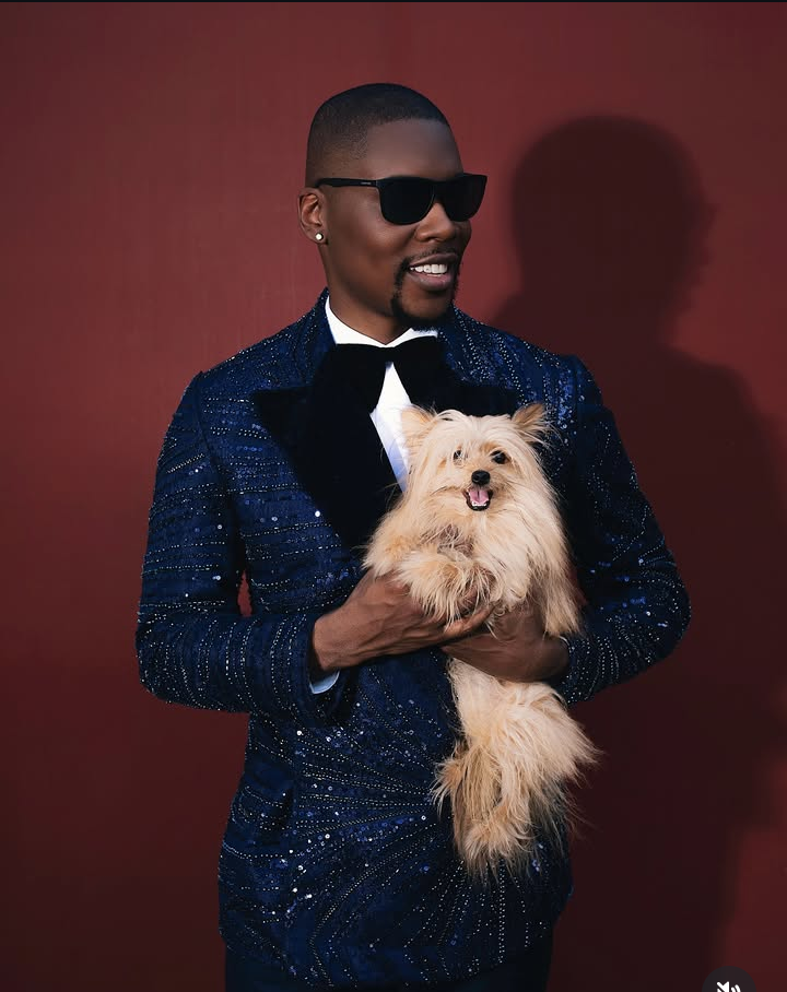 BBNaija star Shaun exudes elegance in a sparkling navy blue suit with beadwork, a crisp white shirt, and a black bow tie, holding a fluffy light brown dog against a rich maroon backdrop.