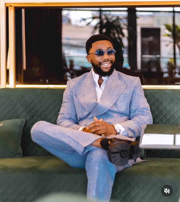 BBNaija star Sooj sits confidently on a green quilted sofa, dressed in a light blue double-breasted suit over a white shirt. The elegant indoor setting features reflective surfaces and lush greenery, enhancing the luxurious and relaxed vibe.