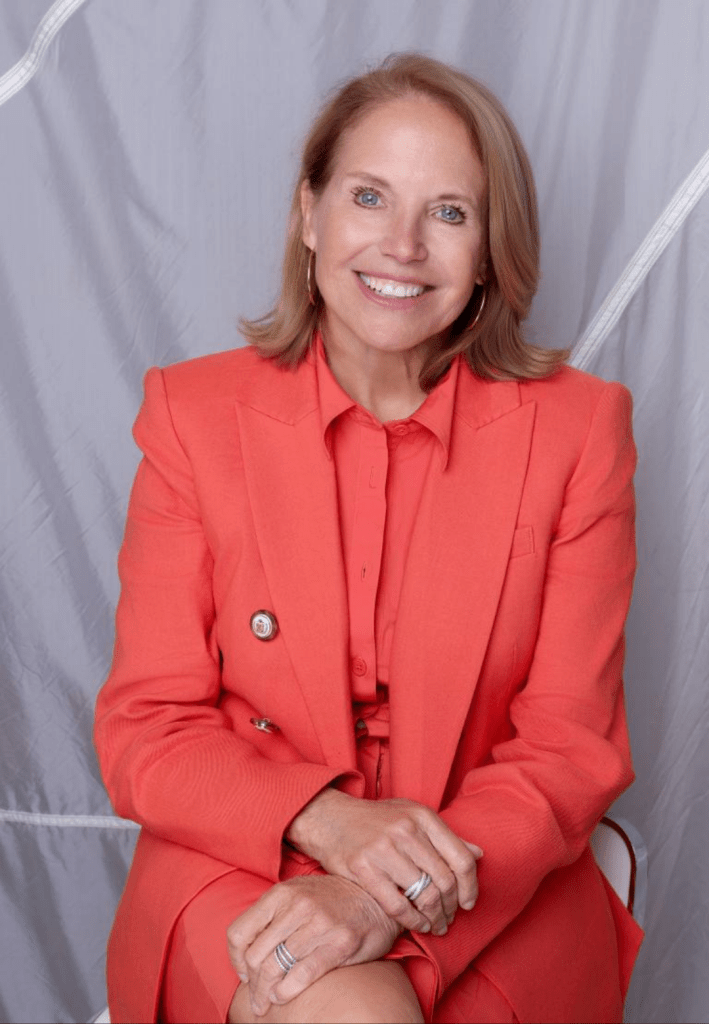 Katie Couric wears a vibrant coral double-breasted suit with silver buttons and a matching collared blouse, her hands adorned with silver rings, set against a neutral gray backdrop.