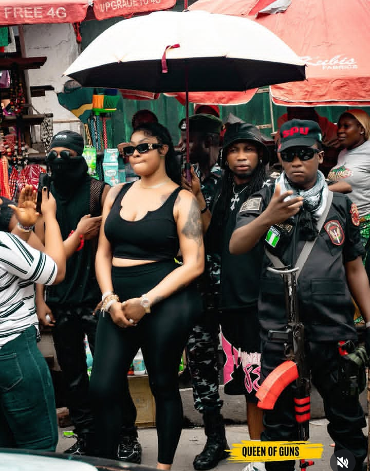 Angela Okorie stands confidently in a bustling outdoor market, dressed in a black sleeveless top and leggings, with tattoos visible on her left arm. Surrounded by security personnel and onlookers, she is shielded by an umbrella held by one of the guards. Red vendor umbrellas and signage promoting mobile services fill the background, while bold text at the bottom reads “QUEEN OF GUNS.”