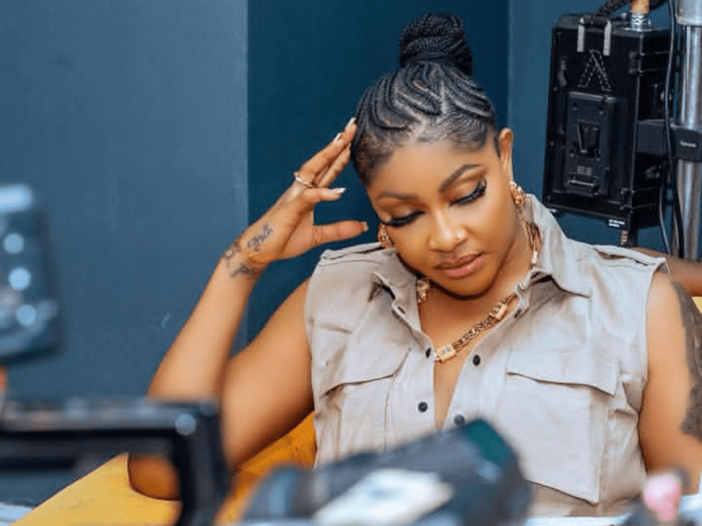 Angela Okorie sits confidently in a film production setting, wearing a sleeveless beige top and gold necklace, with her braided hair styled neatly. Professional camera equipment frames the scene.