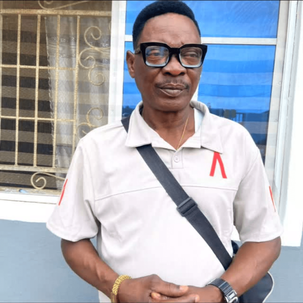 Actor Lalude stands in front of a window with a decorative metal grille, dressed in a light grey polo shirt with red accents, a black crossbody bag, a gold bracelet, and a wristwatch, with hands clasped in front.