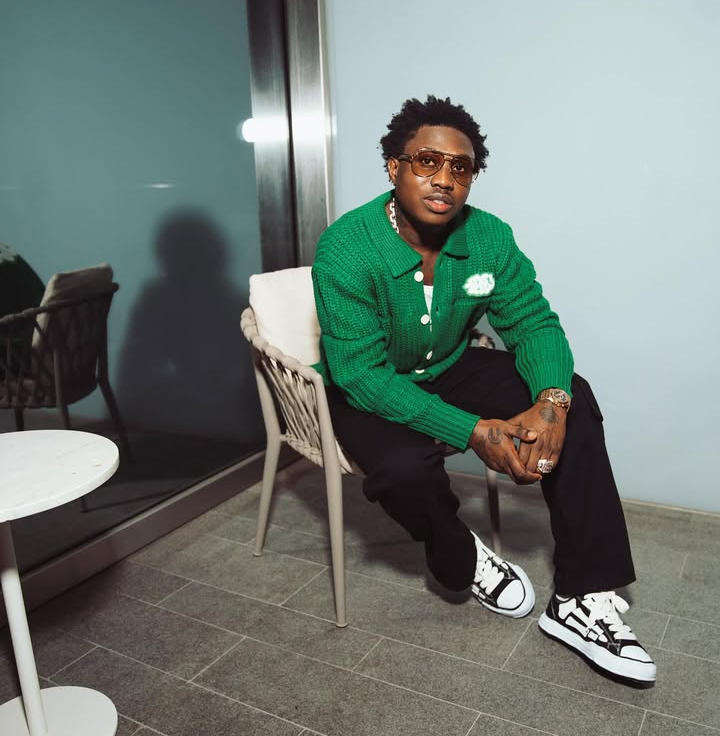 Rapper Zlatan sits confidently in a green cardigan with white buttons and a pocket square, paired with black pants and black-and-white sneakers. He accessorizes with rings and a watch, seated beside a small round table in an indoor setting.