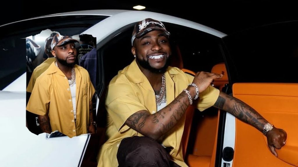 Singer Davido, dressed in a yellow shirt and cap, sits in a white car with an orange interior, accessorized with multiple chains and bracelets, while pointing outward with a confident smile. A reflection of him is visible in the car window.