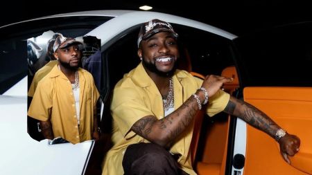 Singer Davido, dressed in a yellow shirt and cap, sits in a white car with an orange interior, accessorized with multiple chains and bracelets, while pointing outward with a confident smile. A reflection of him is visible in the car window.