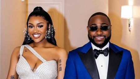 Singer Davido stands beside his wife Chioma, both dressed elegantly. Chioma wears a sparkling, off-shoulder gown with intricate tattoos on her arm and large, ornate earrings, while Davido sports a sharp blue suit with a black bow tie and sunglasses.