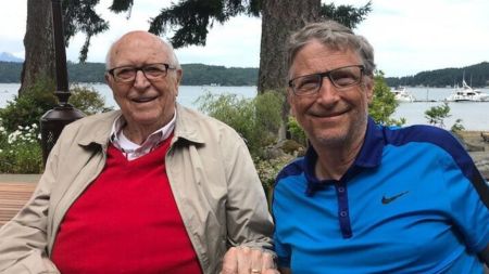 Two men, one in a red sweater and beige jacket and the other in a blue Nike polo shirt, sit side by side outdoors near a lake with boats and trees in the background.
