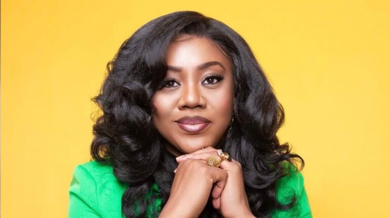 A poised Stella Damasus with long, wavy dark hair, wearing a green top and gold jewelry, stands against a vibrant yellow background with her hands clasped.