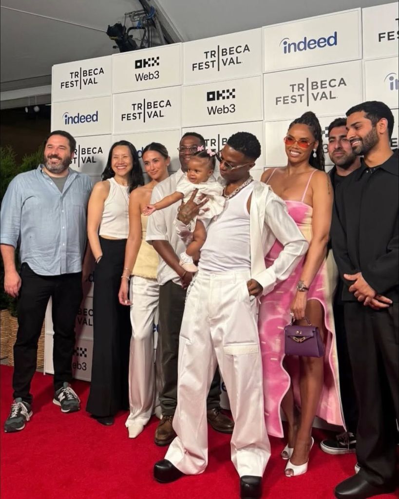 A group of people stands on a red carpet at the Tribeca Festival, with a backdrop featuring logos of Indeed, Web3, and TRIBECA FEST|VAL. The group includes a man in a light blue shirt and dark pants, a woman in a white crop top and black skirt, another woman in a light-colored outfit holding a baby, a man in a white ensemble with sunglasses, a woman in a vibrant pink dress holding a purple handbag, and others in casual and formal attire. The baby, dressed in white, is playfully reaching out, adding a lively touch to the scene.