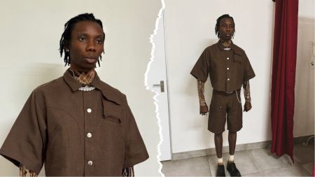 Blaqbonez poses in a brown two-piece outfit with patterned inner sleeves and necklace detail, standing indoors beside a white wall and red curtain.
