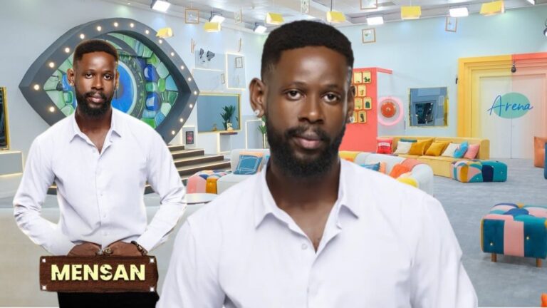 BBNaija Season 10 housemate Mensan posing in a white shirt inside the Big Brother Naija house, with the iconic eye logo and colourful lounge area in the background.