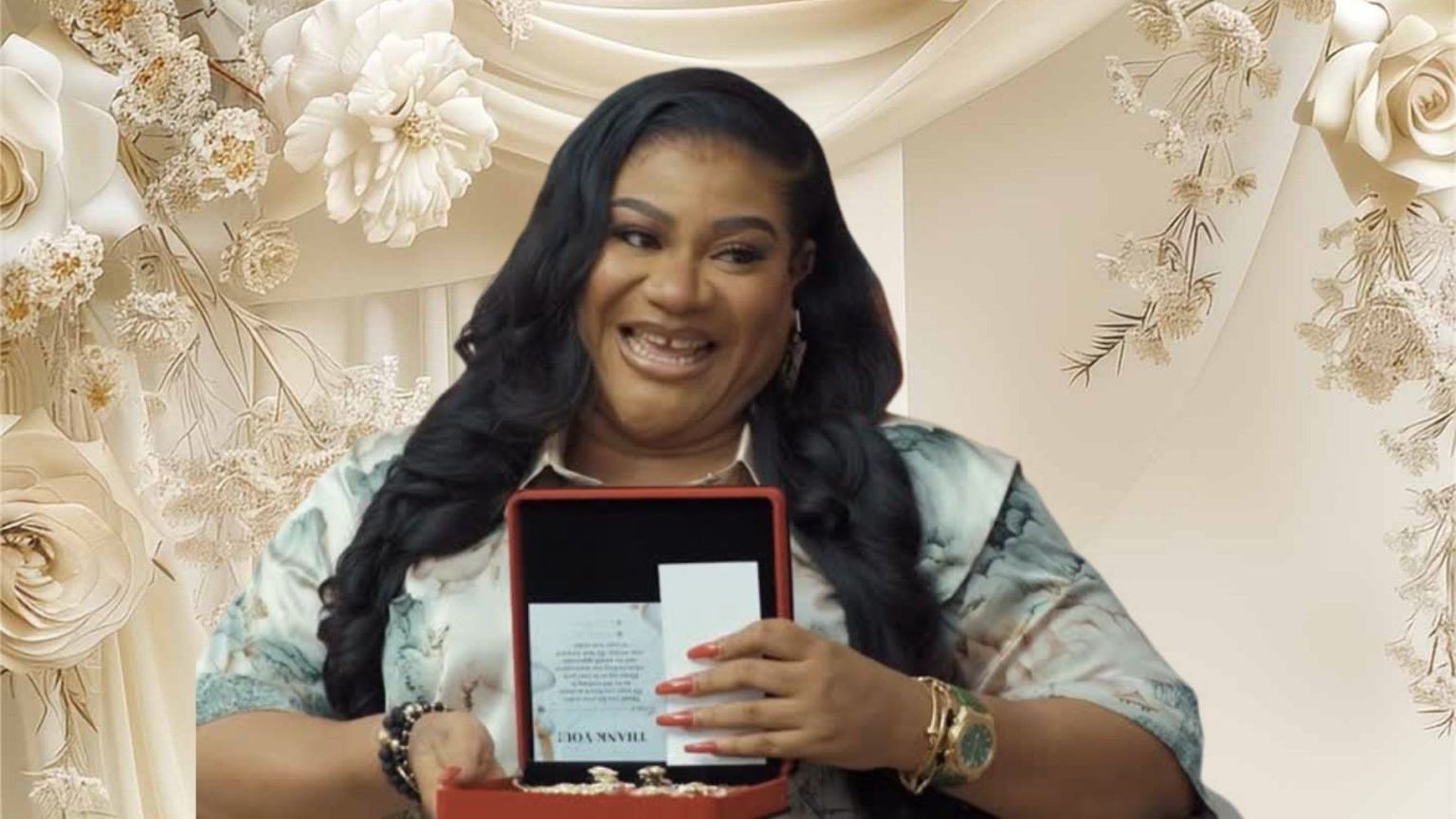Actress Nkechi Blessing smiling while holding an open red gift box with a thank-you card inside. She is dressed in a patterned outfit with gold jewellery and styled wavy hair, posing against a floral backdrop.