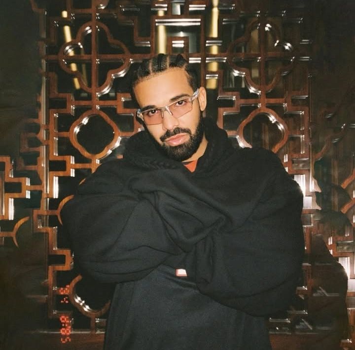 Rapper Drake stands with arms crossed in a large black hoodie against an ornate wooden lattice backdrop, blending casual style with a touch of artistic elegance.