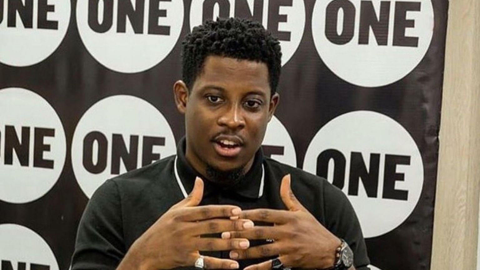 BBNaija star Seyi Awolowo speaking during a media engagement, dressed in a black polo shirt with a backdrop featuring repeated "ONE" logos.