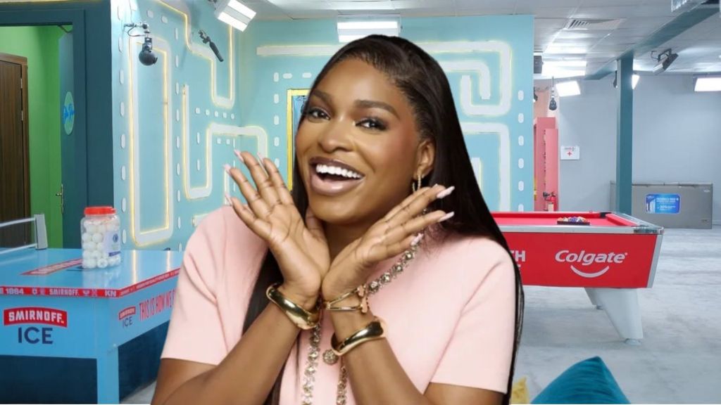 BBNaija Season 10 housemate Thelma Lawson smiling brightly in a pink outfit inside the Big Brother Naija house lounge, with a pool table and branded game area visible in the background.