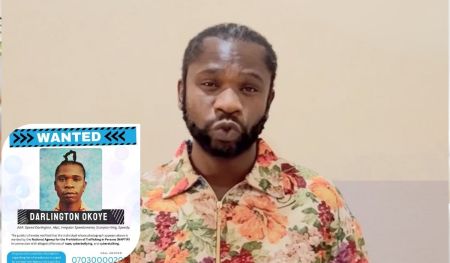 Rapper Speed Darlington, dressed in a vibrant floral shirt, stands beside a “WANTED” poster bearing his name and aliases, issued by NAPTIP in connection with serious allegations.