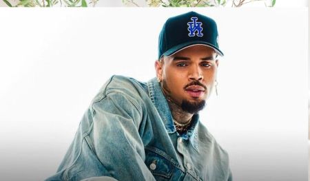 Chris Brown wears a denim jacket and a dark blue cap with a white and blue logo, accessorized with multiple gold chains and earrings, set against a white backdrop with green plants overhead.