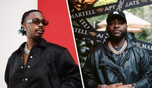 Young Jonn stands confidently in a black jacket adorned with silver buttons and a chunky chain necklace, while Davido sports a black leather Puma hoodie and matching chain, set against a backdrop featuring a Martell promotional banner.