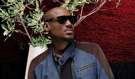 Singer 2baba wears a denim jacket with brown suede shoulder patches over a black shirt, standing before a red and black textured backdrop with a white vase of green leafy branches beside him.