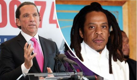Tony Buzbee, in a dark suit with a pink tie, speaks at a podium with bold red numbers in the background, while Jay Z, wearing a white shirt and sporting long dreadlocks, stands beside him.