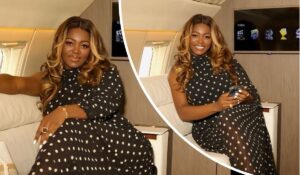 BBNaija star Ka3na exudes elegance aboard a private jet, dressed in a chic black polka dot dress with loose curls framing her face. She holds a smartphone, surrounded by a sleek interior and an entertainment screen, capturing a moment of luxury and style.