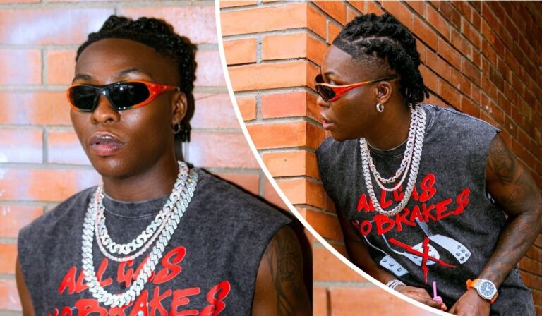 Reekado Banks poses in a sleeveless black shirt with bold red text reading "ALL GAS NO BRAKES" and a graphic of crossed brake pedals. He sports layered silver chains, a watch, and earrings, set against a brick wall that adds an urban edge.