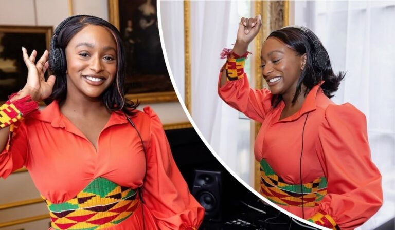 DJ Cuppy wears a vibrant orange dress with puffed sleeves and a patterned belt, holding headphones in one hand and raising the other mid-performance. The elegant setting features framed artwork and draped curtains, enhancing the lively yet refined atmosphere.