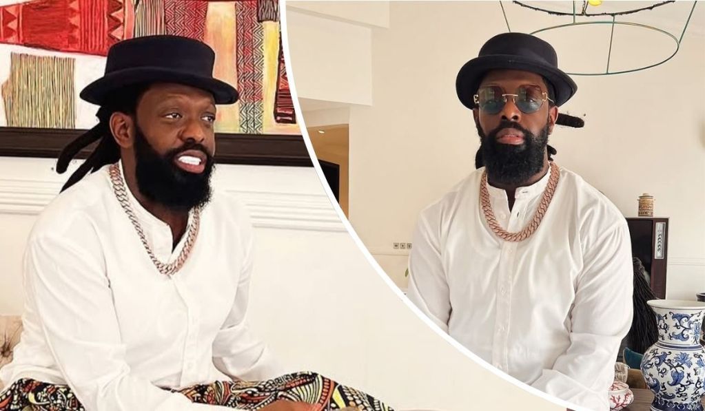 Singer Timaya wears a white traditional outfit with a bold chain necklace and black hat, set against a vibrant backdrop of patterned artwork and decorative pieces, including a blue and white vase. The stylish indoor setting enhances the contemporary vibe.