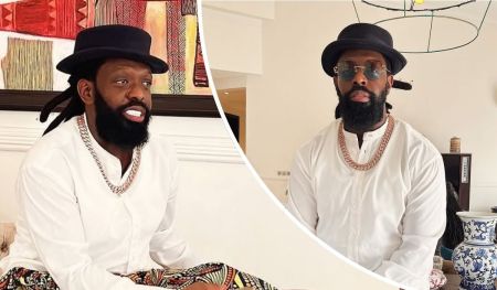 Singer Timaya wears a white traditional outfit with a bold chain necklace and black hat, set against a vibrant backdrop of patterned artwork and decorative pieces, including a blue and white vase. The stylish indoor setting enhances the contemporary vibe.