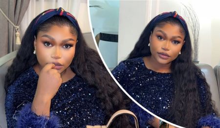 Actress Ruth Kadiri Ezerika poses in a dark blue sequined top with fluffy cuffs, pearl earrings, and a colorful headband. Her long, wavy hair flows over her shoulders as she sits in a chic, modern room featuring a grey curtain, beige armchair, and a beige handbag placed in front of her.