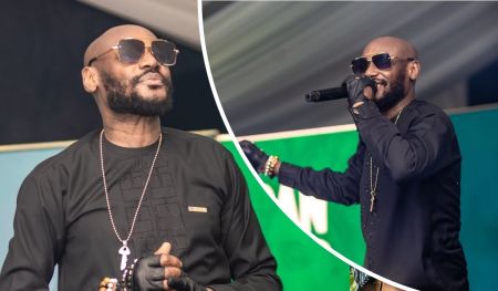 Singer 2baba performs on stage in a patterned black outfit, accessorized with black gloves and a key pendant necklace, passionately engaging the audience with a microphone in hand against a green backdrop.