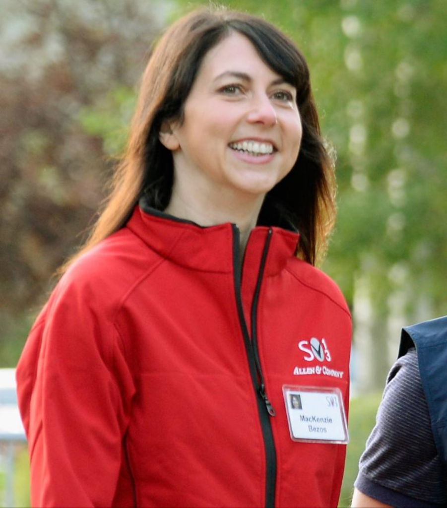 MacKenzie Scott wears a red Allen & Company jacket with a name tag labeled "MacKenzie Bezos," standing outdoors in a natural setting.