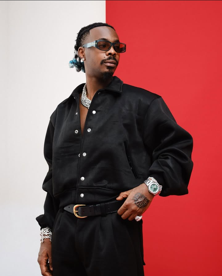 Young Jonn poses in a black button-up jacket and matching pants, accessorized with a chunky silver necklace, bracelet, and watch, his hair styled with blue highlights against a bold red-and-white backdrop.