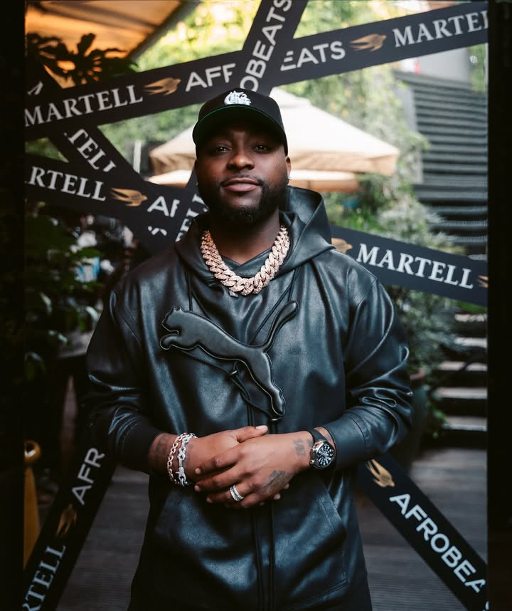 Davido wears a black leather Puma hoodie paired with a thick gold chain, black wristwatch, and layered bracelets, standing before Martell and Afrobeats promotional banners at an outdoor event.