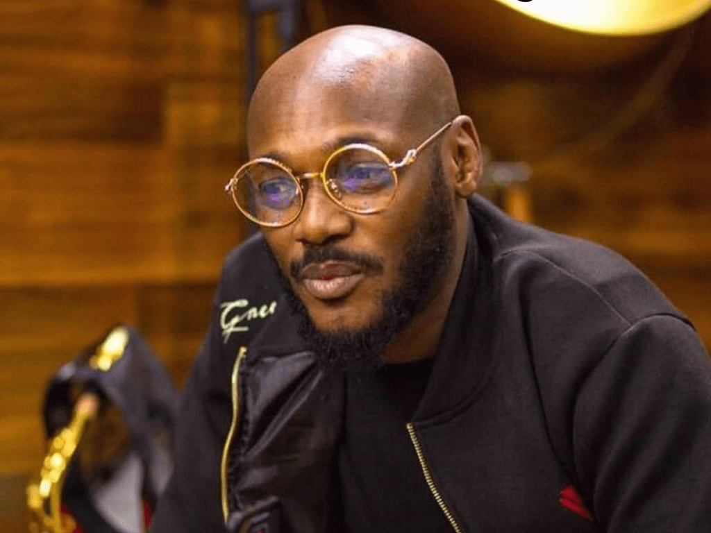 Nigerian singer 2baba, dressed in a black zip-up jacket, holds a mobile phone against a backdrop of wooden paneling and a golden object, with the "AFROMIXX" logo in the corner.