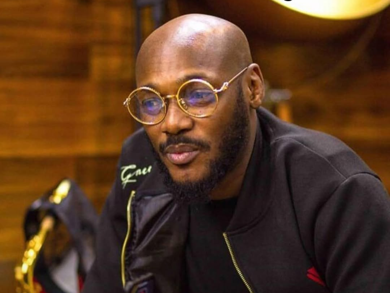 Nigerian singer 2baba, dressed in a black zip-up jacket, holds a mobile phone against a backdrop of wooden paneling and a golden object, with the "AFROMIXX" logo in the corner.