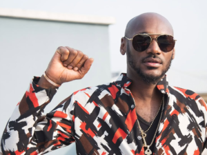 Singer 2baba wears a vibrant, patterned shirt with abstract designs in black, white, red, and brown, accessorized with layered necklaces and a bracelet. He raises his right fist in a gesture of solidarity, set against a light-colored building and clear sky.