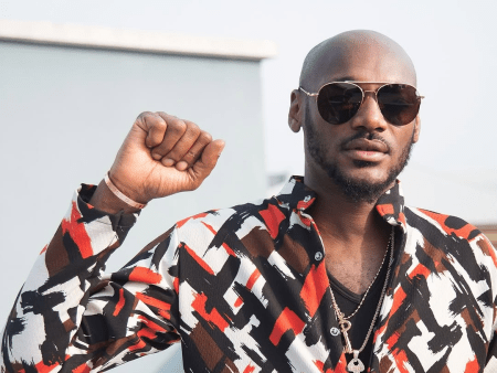 Singer 2baba wears a vibrant, patterned shirt with abstract designs in black, white, red, and brown, accessorized with layered necklaces and a bracelet. He raises his right fist in a gesture of solidarity, set against a light-colored building and clear sky.