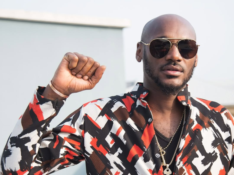 Singer 2baba wears a vibrant, patterned shirt with abstract designs in black, white, red, and brown, accessorized with layered necklaces and a bracelet. He raises his right fist in a gesture of solidarity, set against a light-colored building and clear sky.