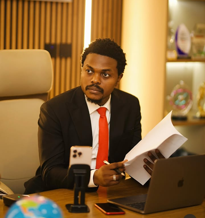 Bitcoin mogul Blord sits confidently at his office desk in a black suit and red tie, holding a document and pen. Behind him, shelves lined with awards and trophies highlight his professional achievements.