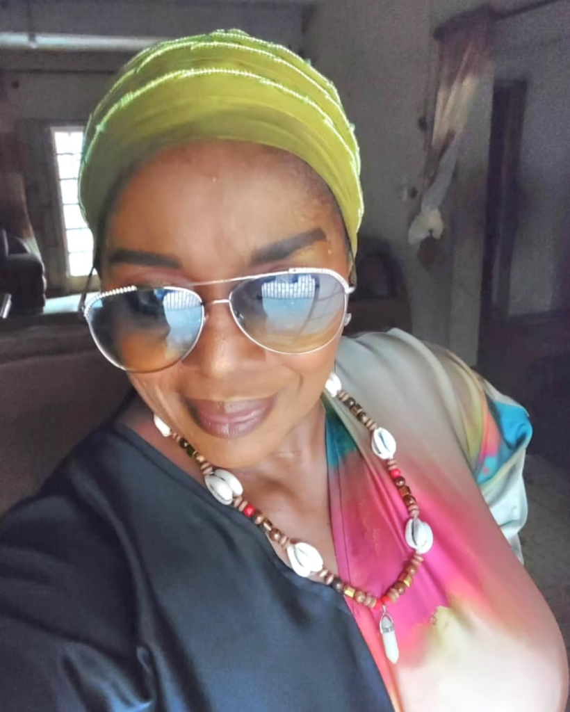 Actress Rita Edochie wears a vibrant multi-colored outfit adorned with a beaded and cowrie shell necklace, complemented by a bright yellow headwrap, standing in a dimly lit room with a window and furniture in the background.