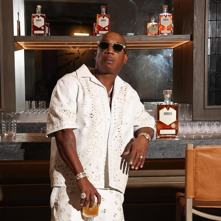 Rap star Ja Rule stands at a sleek bar in a patterned white outfit, holding a drink with bottles of Amber liquor displayed behind him, creating a stylish and upscale vibe.