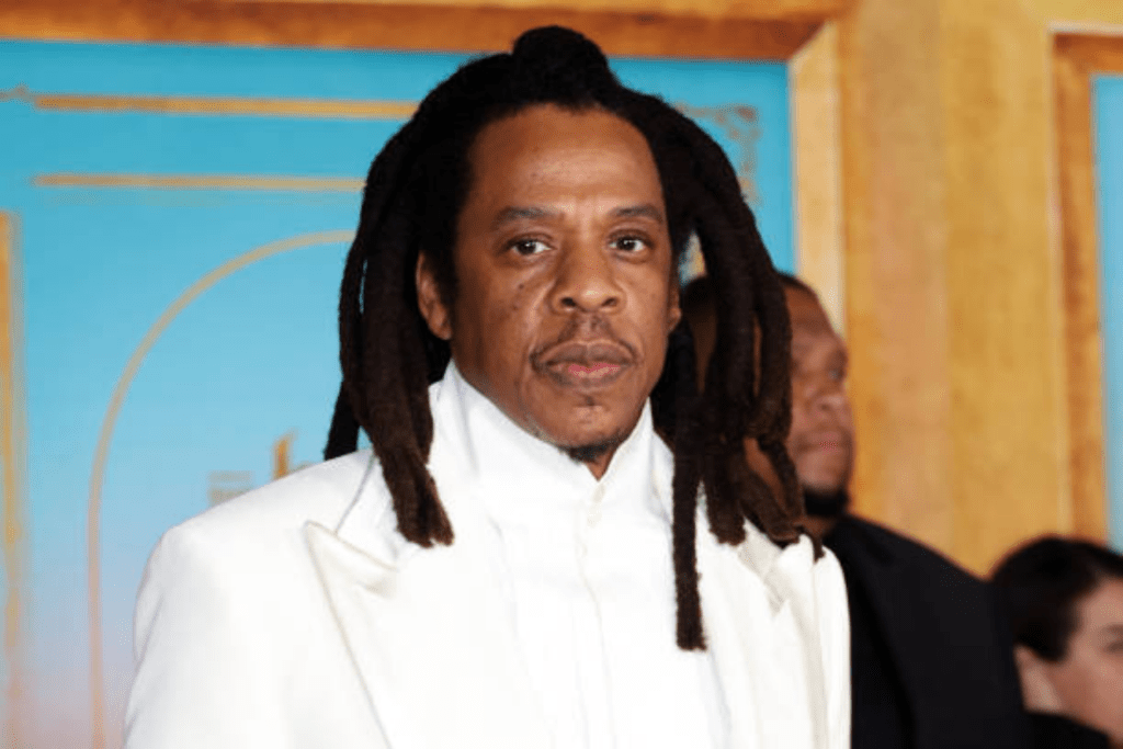 Jay Z wearing a white suit with a high-collared shirt, his natural textured hair styled in freeform locs, standing against a blue and gold patterned backdrop