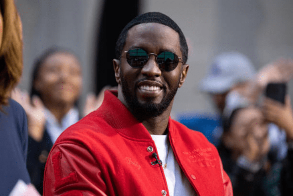 Rapper Diddy wears a red embroidered jacket over a white shirt with a microphone clipped to his chest, surrounded by onlookers capturing the moment at what appears to be a public or media event.