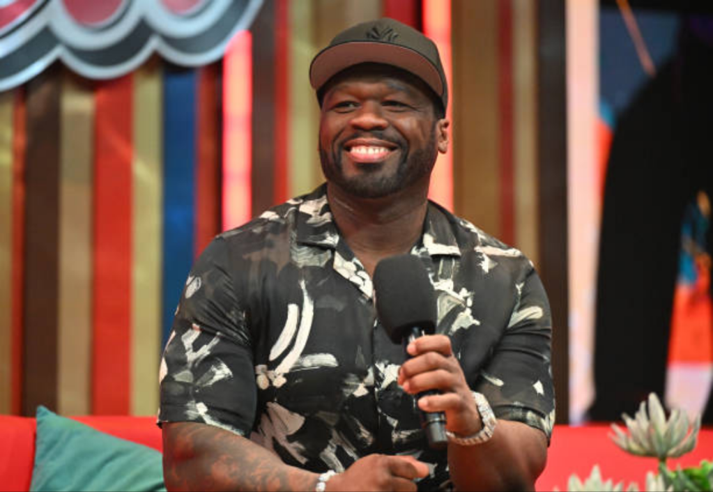 50 Cent sits on a red couch, holding a microphone and wearing a black and white patterned shirt with a cap. Behind him, colorful vertical stripes and lush plants add vibrancy to the setting.