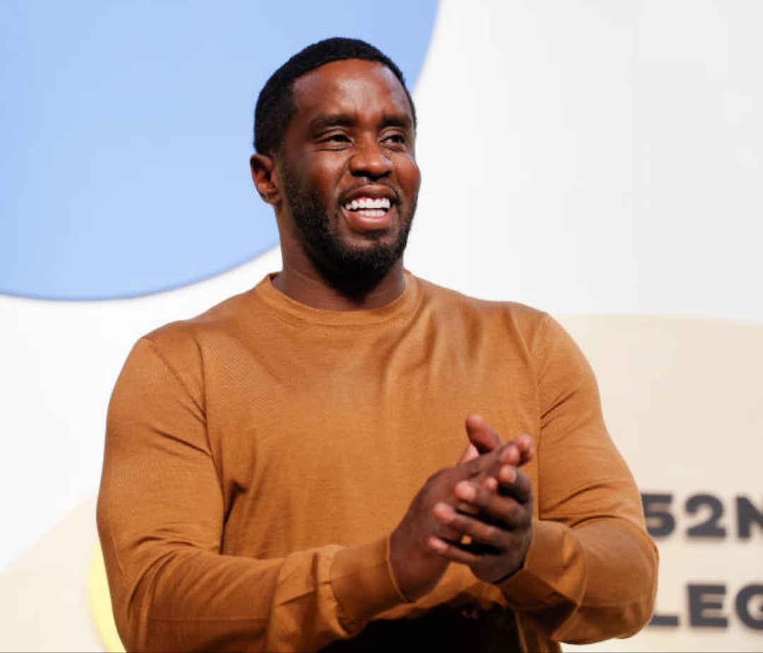 Diddy wears a brown long-sleeve shirt and claps his hands in front of a backdrop featuring a blue circular design and partial text reading “52N LEG.”